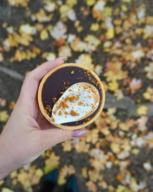 
                  
                    Load image into Gallery viewer, Hand holding a small round dessert with a dark chocolate ganache smores and white marshmallow topping, against a blurred autumn leaf background.
                  
                